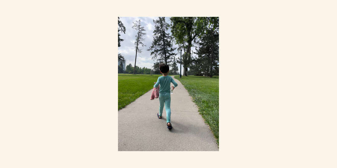 A young child in teal pajamas walks down a wide sidewalk through a green park, holding a pink stuffed animal and a book, with tall trees framing the path ahead.